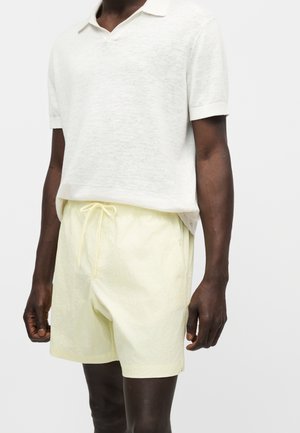 Mężczyzna ubrany w lekką białą koszulkę polo z krótkim rękawem i bladoróżowe spodenki ze sznurkiem na jednolitym tle.