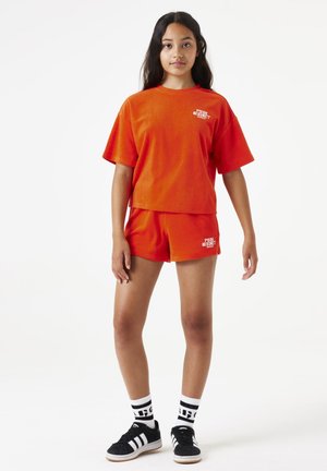 Top de manga corta de toalla de rizo naranja y pantalones cortos a juego. El top presenta el texto blanco "Pink Sunset"; el modelo lleva zapatos negros y calcetines a rayas.