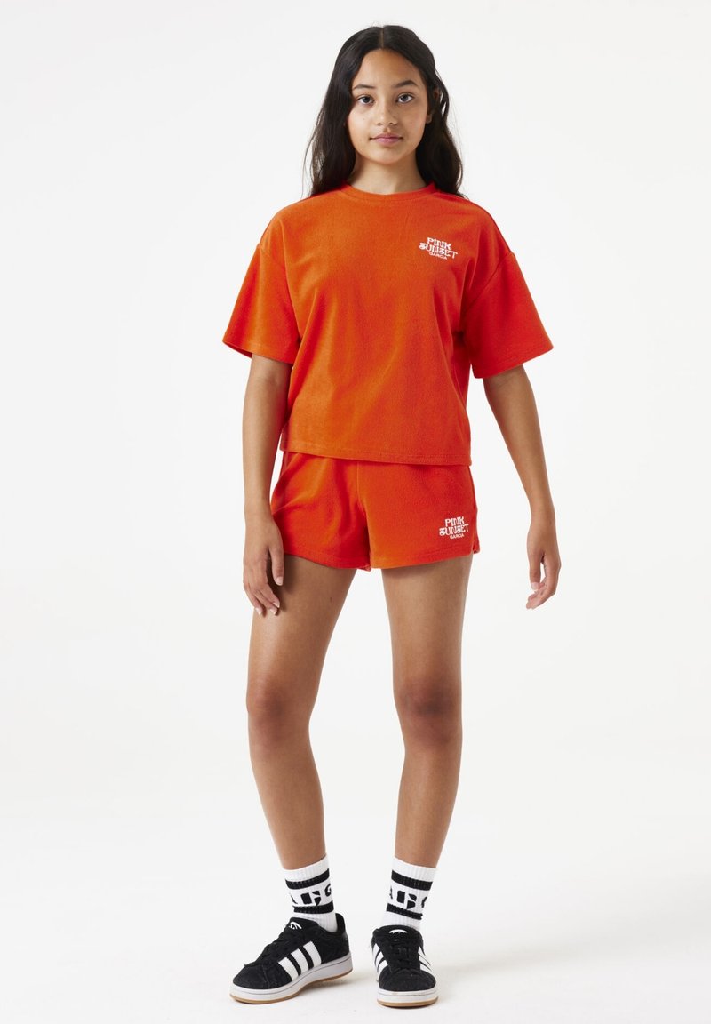Oranje badstof top met korte mouwen en bijpassende shorts. De top heeft witte tekst "Pink Sunset"; het model draagt zwarte schoenen en gestreepte sokken.