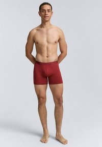 Rote Baumwoll-Boxershorts mit einem strukturierten Bund, figurbetontem Design und glattem Finish, getragen von einem männlichen Modell, das barfuß steht.