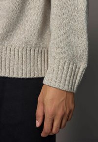 Pull en tricot beige clair, montrant un tissu texturé et des poignets côtelés. Main posée sur un tissu foncé, mettant en avant le contraste de couleurs et les détails.