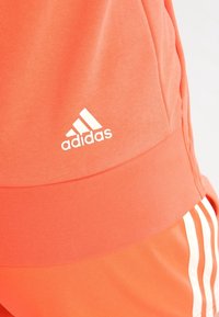 Shorts de sport Adidas orange avec une texture lisse. Dotés d'un logo blanc et de rayures blanches verticales le long des côtés.