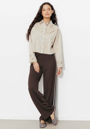 Femme debout portant une chemise beige courte à boutons avec des détails découpés, un pantalon large foncé à rayures latérales, et des ballerines imprimé léopard.