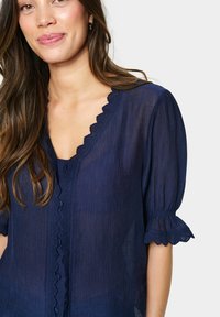 Blusa blu navy con scollatura e orlo scallop. Realizzata in tessuto leggero con un motivo testurizzato, presenta maniche corte a palloncino.