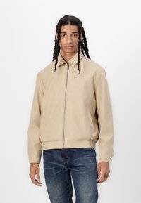 Veste bomber beige avec une fermeture éclair sur le devant, des poches latérales et des poignets côtelés. Fabriquée dans un tissu lisse avec un design minimaliste.