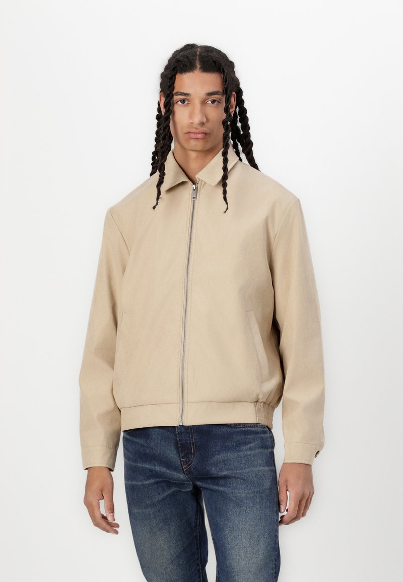 Veste bomber beige avec une fermeture éclair sur le devant, des poches latérales et des poignets côtelés. Fabriquée dans un tissu lisse avec un design minimaliste.