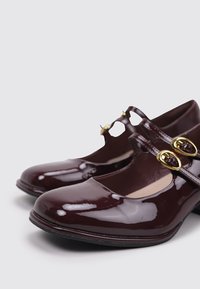 Zapatos Mary Jane en charol burdeos brillante. Presentan dos hebillas doradas y un diseño de punta redondeada. Tacón bajo con una textura suave y brillante.