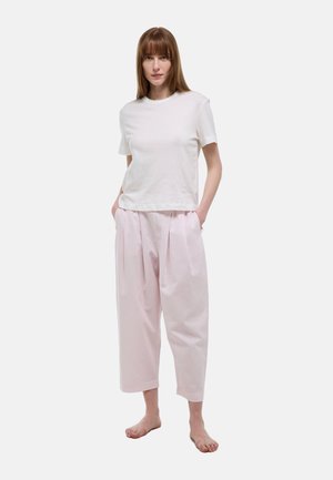 Femme aux cheveux bruns raides, debout pieds nus, portant un t-shirt blanc uni à manches courtes et un pantalon ample rose clair à jambes larges et raccourci.