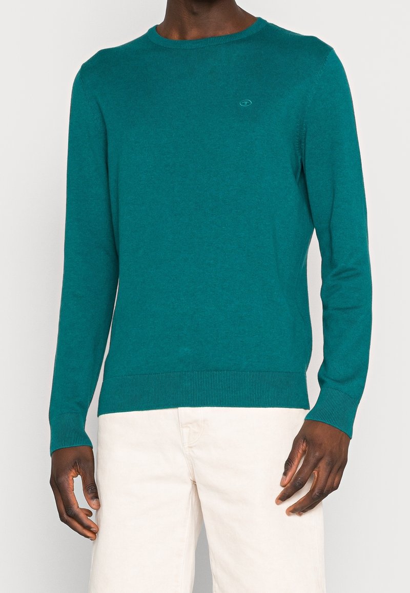 Homme portant un pull à manches longues couleur sarcelle et un pantalon écru, debout devant un fond clair uni avec une posture détendue.