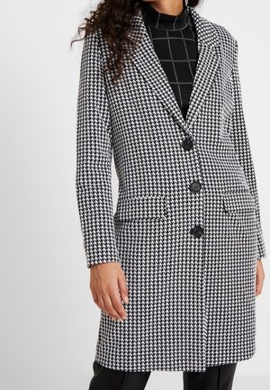 Femme portant un manteau boutonné à motif pied-de-poule noir et blanc sur un haut noir avec des lignes de quadrillage blanches.