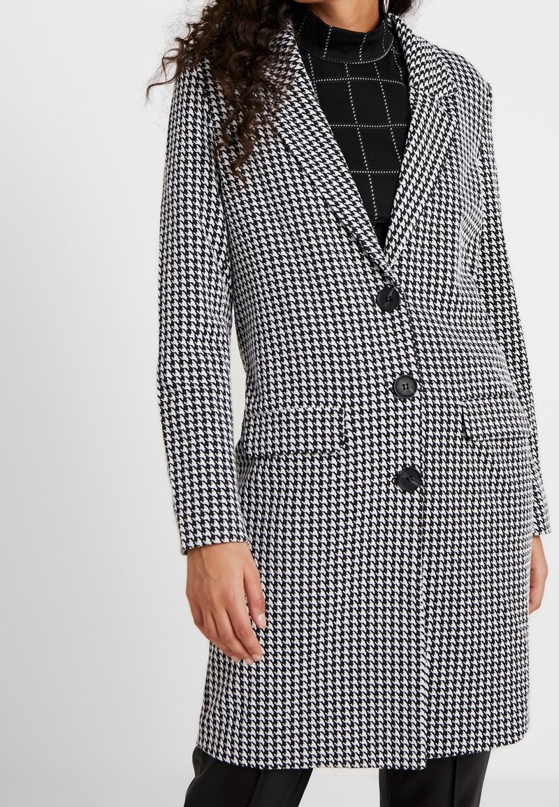 Femme portant un manteau boutonné à motif pied-de-poule noir et blanc sur un haut noir avec des lignes de quadrillage blanches.