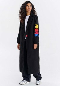 Kobieta z długimi kręconymi włosami, ubrana w długi czarny płaszcz z kolorowym napisem "MORE" na rękawie, białą koszulę, ciemne spodnie i białe trampki.