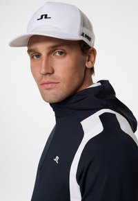 Vit keps med meshnack och svart logotyp. Modellen bär en marinblå zip-jacka med vita accentlinjer på ärmarna. Enkel, sportig design.