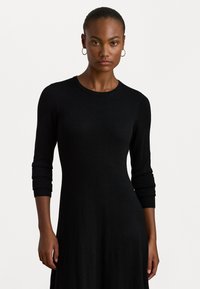 Vestido de punto negro con mangas de tres cuartos, cuello redondo y un cuerpo ajustado que se despliega en una falda fluida. Textura suave.