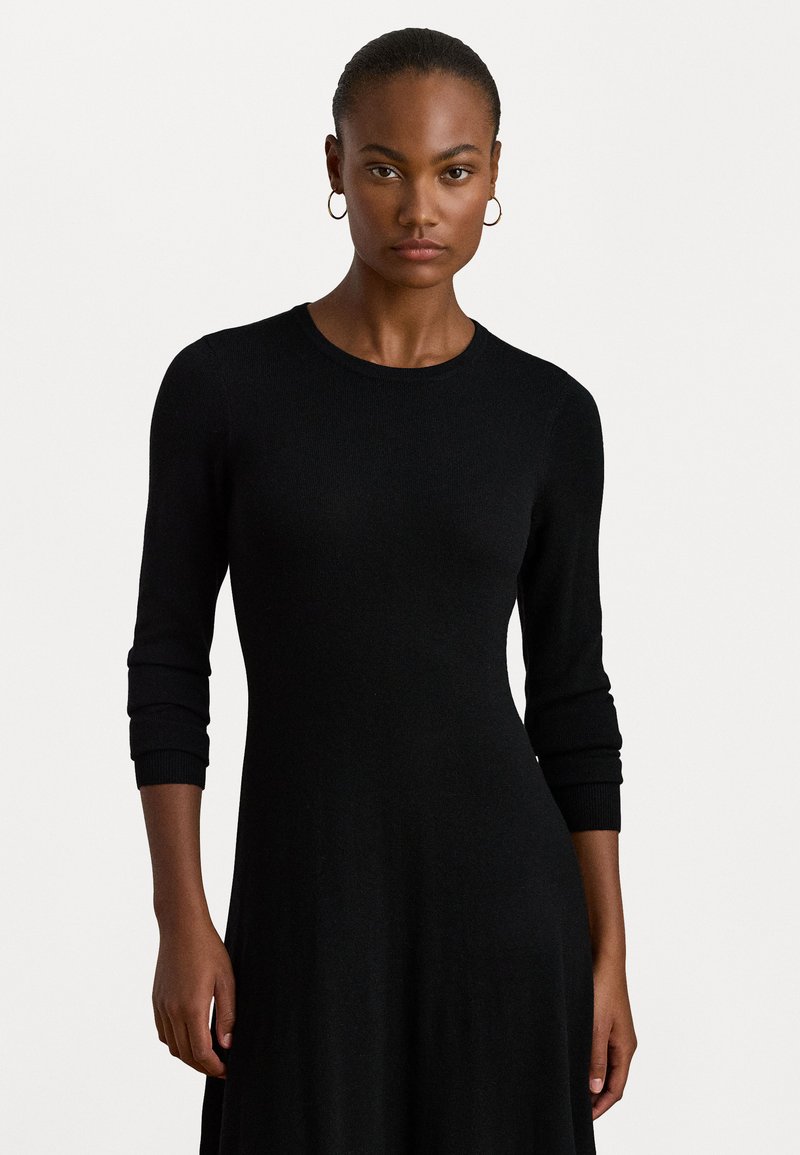 Vestido de punto negro con mangas de tres cuartos, cuello redondo y un cuerpo ajustado que se despliega en una falda fluida. Textura suave.