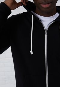 Sudadera con cremallera negra y cordón blanco. Cuenta con una cremallera de metal y un bolsillo frontal. Material de mezcla de algodón, textura suave, diseño de peso medio.