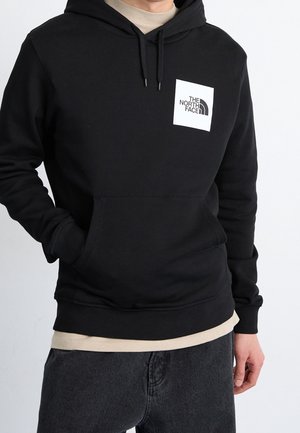 Person iført en sort pullover-hoodie med "The North Face"-logo på brystet, beige skjorte under, og mørkegrå bukser.