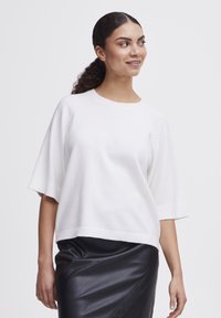 Witte gebreide pullover met korte mouwen, ronde hals en geribde zoom, gecombineerd met een aansluitende zwarte leren rok. Glad textuur.