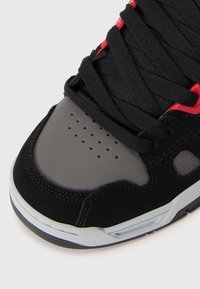 Close-up of black and gray sneaker toe with black laces, perforations on gray area, and red lace eyelets on a white background.