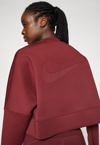 Bordový zkrácený sweatshirt vyrobený z hladké tkaniny, s tónovým logem Nike na zadní straně, širokými manžetami a volným střihem.
