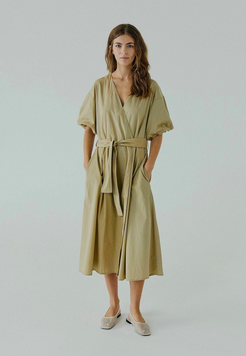 Robe portefeuille beige avec des manches courtes bouffantes, une taille ceinturée et des poches latérales. Tissu lisse avec une silhouette fluide, portée avec des chaussures à bout ouvert.