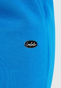 Helles blaues Gewebe mit einer glatten Textur, versehen mit einem schwarzen ovalen Logo-Patch, auf dem "Smilodox" in weißer Schreibschrift steht.