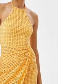 Gul halterneck-klänning i texturerad tyg med utskurna mönster. Den har en samlad detalj på sidan för en draperad look.