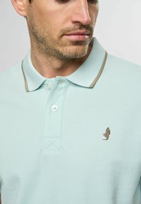 Ljusblå pikétröja i texturerad tyg, med en kontrasterande beige krage och en liten broderad figur på vänster bröst.