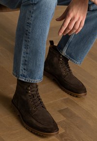 Stivali ankle in suede marrone con punta arrotondata, lacci e occhielli in metallo; superficie texturizzata e suola in gomma, abbinati a jeans di jeans azzurri chiari.