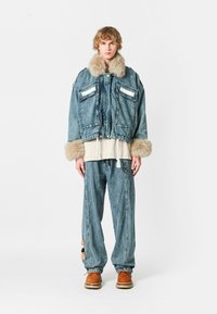 Chaqueta de mezclilla con cuello y puños de piel, dos bolsillos frontales, combinada con pantalones de mezclilla holgados con un parche decorativo y calzado marrón.