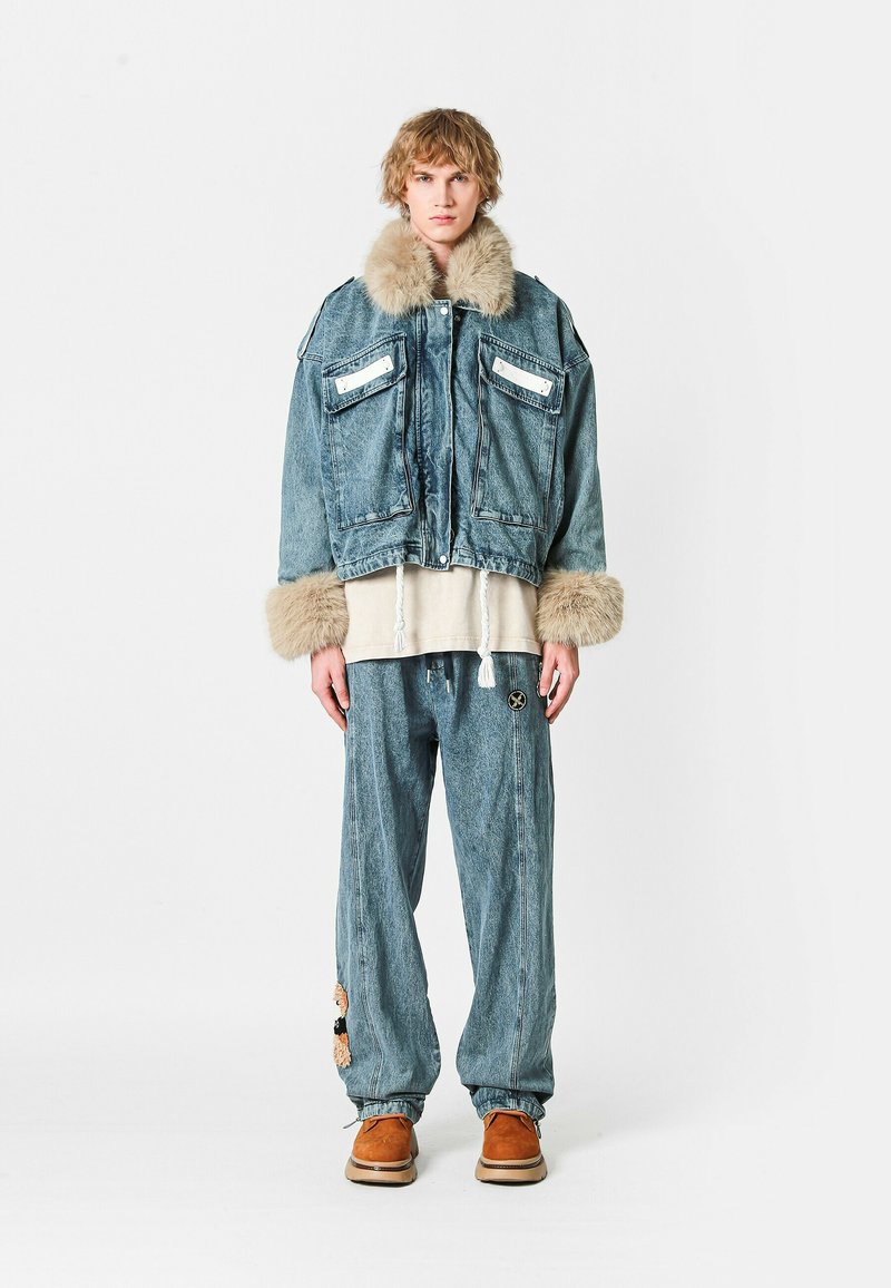 Chaqueta de mezclilla con cuello y puños de piel, dos bolsillos frontales, combinada con pantalones de mezclilla holgados con un parche decorativo y calzado marrón.