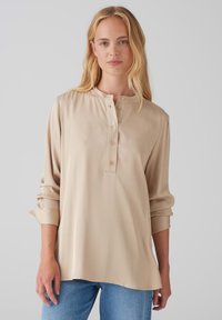 Beige Langarmbluse mit Henley-Kragen, ausgestattet mit vier Knöpfen auf der Vorderseite. Glatter, leichter Stoff mit aufgerollten Manschetten.