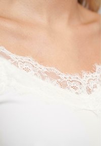Witte stof top met kantafwerking en delicate bloemenpatronen bij de halslijn, die een zachte textuur en ingewikkelde details toont.