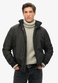 Chaqueta bomber negra con cuello alto y cremallera, llevada sobre un suéter de punto gris claro. Tela texturizada con puños acanalados, diseño ajustado.