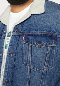 Man i blå denimjacka med vit sherpa-krage över vit t-shirt med grafiskt tryck, avkortad vid bröstet och halsen.