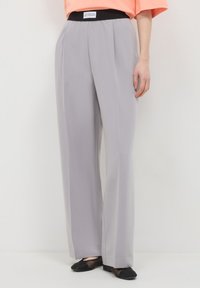 Pantalons droits gris clair avec une étiquette de ceinture noire portant l'inscription "MOINS DE PERFECTION, PLUS D'AUTHENTICITÉ", portés avec des ballerines noires et un top corail.