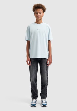 Jong persoon met krullend haar, gekleed in een lichtblauw oversized T-shirt, vervaagde zwarte jeans en witte sneakers met blauwe details, staand rechtop.