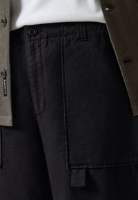 Black cotton cargo pants with side pockets, featuring a visible waistband and button closure. Textured olive-green shirt partially visible.