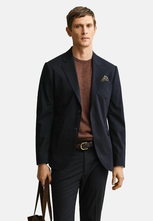 Homme portant un blazer sombre avec un mouchoir de poche fleuri, un pull marron, un pantalon foncé, tenant un sac fourre-tout en cuir marron sur fond blanc.