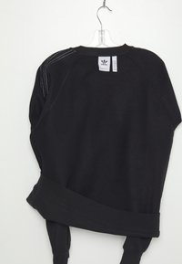 Svart Adidas sweatshirt hängande på en klar plastgalge, visar baksidan med storleks- och varumärkesetiketter, samt vita ränder på axeln.