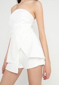 Strapless white romper with a fitted bodice, layered asymmetrical hem, and shorts underneath. Made of smooth, lightweight fabric.