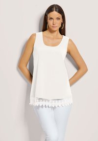 Weißes ärmelloses Oberteil mit quadratischem Ausschnitt und Spitzenbesatz am Saum, kombiniert mit hellblauen Hosen. Glatte Textur, lockere Passform.