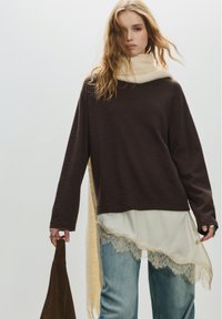 Barna, hosszú ujjú felső texturált felülettel, krémszínű felső rétegként, amely csipkés szegéllyel díszített. Világoskék farmerrel és sötétbarna táskával párosítva.