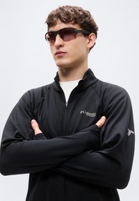 Μαύρο μπουφάν με φερμουάρ και μακριά μανίκια, με υφή, με το λογότυπο της Columbia. Φοράει σκούρα γυαλιά ηλίου, με τα χέρια σταυρωμένα.