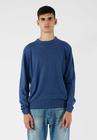 Niebieski sweter z dzianiny z długimi rękawami, okrągłym dekoltem, �ściągaczami przy mankietach i u dołu. Noszony z jasnoniebieskimi dżinsami, na białym tle.
