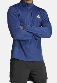 Námořnická modrá sportovní košile s dlouhým rukávem a límcem na půlměsíc, s bílým logem na levé hrudi. Tkanina vypadá hladce a lehce.