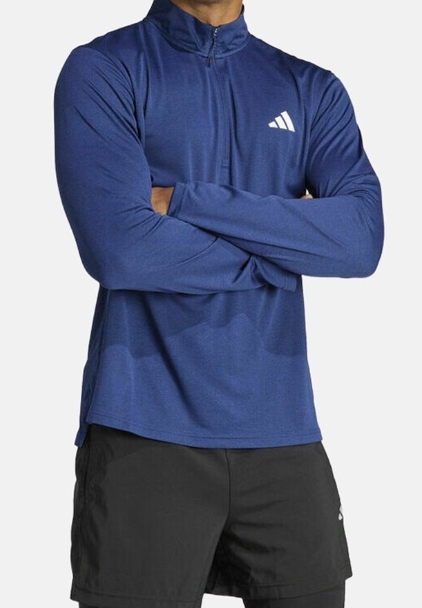 Námořnická modrá sportovní košile s dlouhým rukávem a límcem na půlměsíc, s bílým logem na levé hrudi. Tkanina vypadá hladce a lehce.