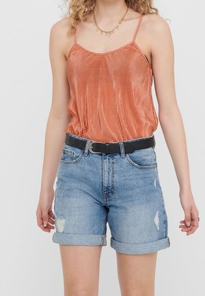 Vrouw draagt een oranje mouwloos topje, ingestopt in opgerolde lichtblauwe denimshorts met een zwarte riem en een gouden ketting.