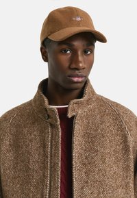 Brown wool cap with embroidered logo, paired with a textured herringbone brown jacket and a maroon cable-knit sweater underneath.