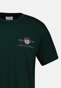 Dark green cotton t-shirt with round neck. Features a printed logo in white and red with "1949" and "AMERICAN SPORTSWEAR" text. Short sleeves.